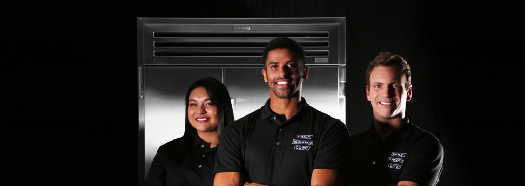 Three people wearing black collared shirts stand in front of a large stainless steel Wolf kitchen appliance, smiling at the camera against a dark background.