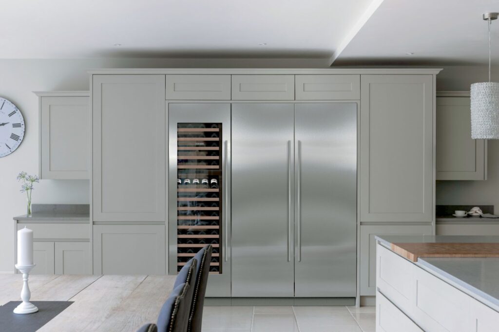 Modern kitchen with light gray cabinets, a built-in stainless steel fridge featuring an ice maker, a wine cooler, and minimalistic decor including a wall clock and candlesticks on the dining table.