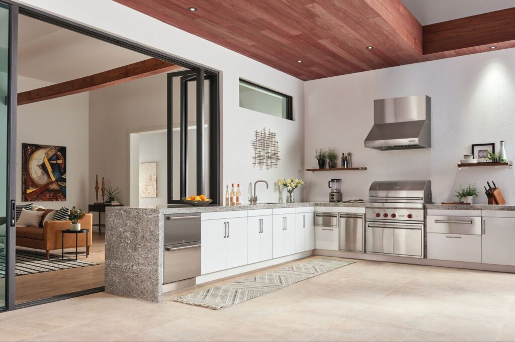 Modern indoor-outdoor kitchen with stainless steel appliances—including a built-in ice maker—white cabinets, open shelving, and sliding glass doors leading to a living area.