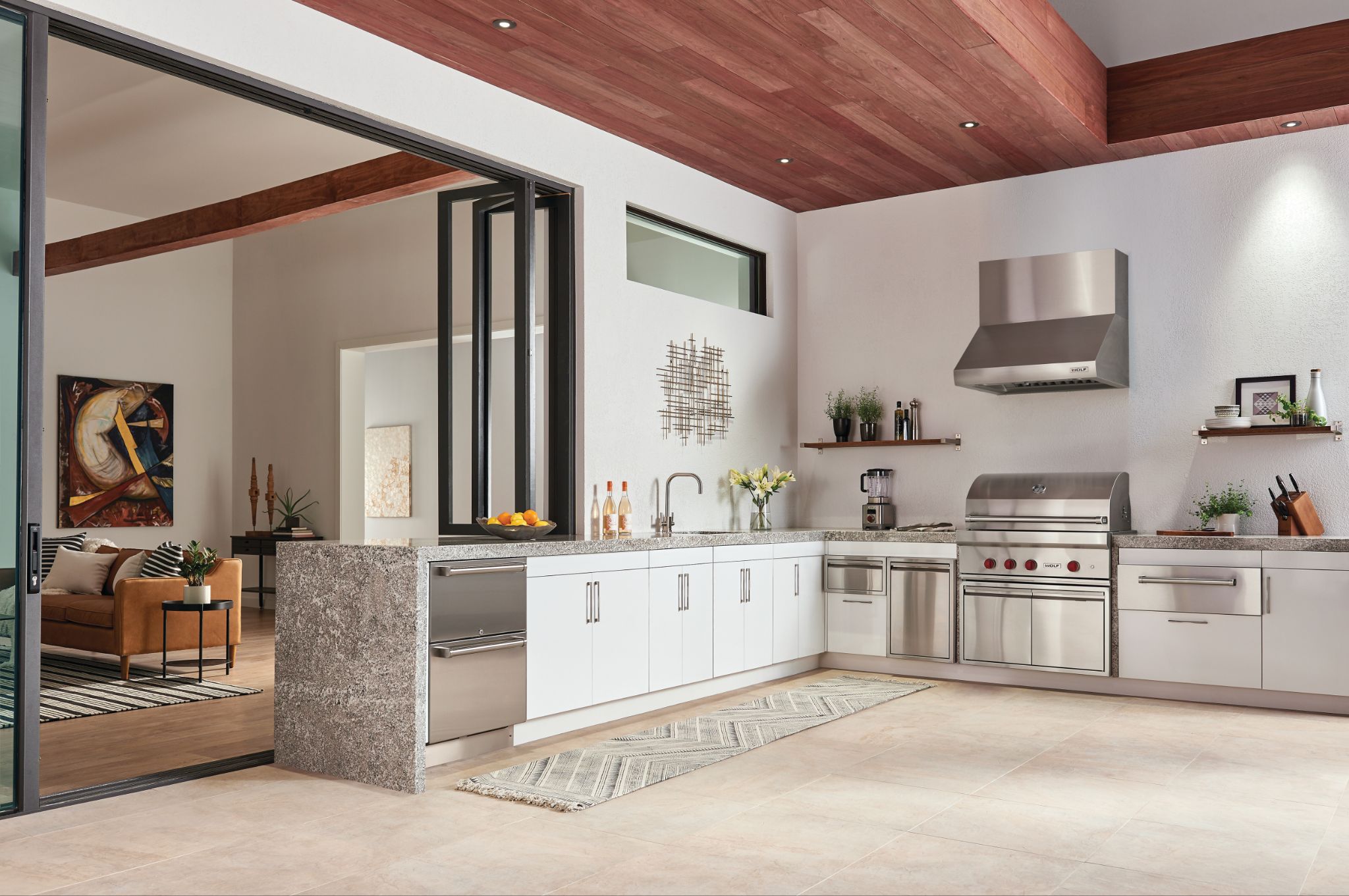 Modern indoor-outdoor kitchen with stainless steel appliances—including a built-in ice maker—white cabinets, open shelving, and sliding glass doors leading to a living area.