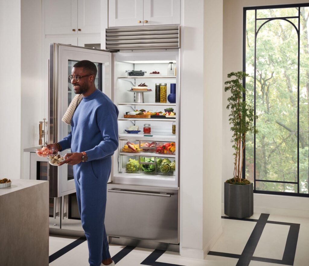 A man in blue loungewear stands in a modern kitchen holding a bowl, with the refrigerator door open, revealing organized food items inside.