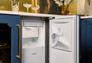Open under-counter ice maker filled with ice, with a scoop hanging on the door, situated in a blue cabinet kitchen with gold accents.