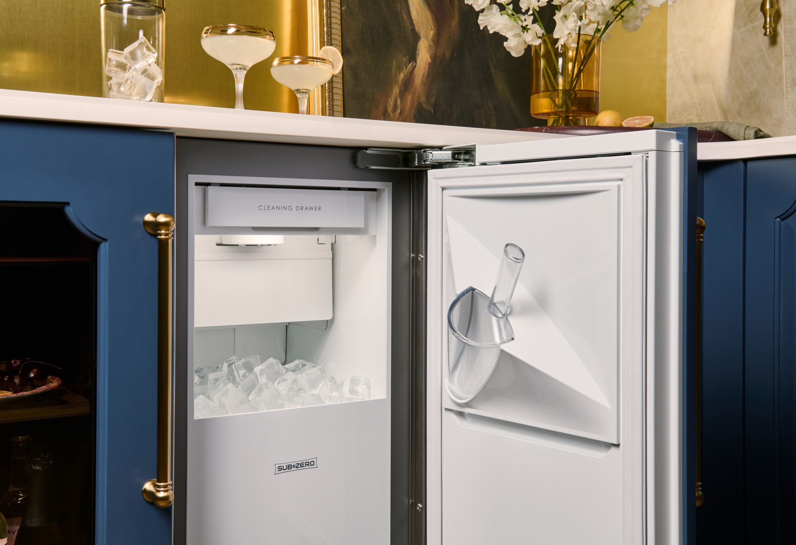 Open under-counter ice maker filled with ice, with a scoop hanging on the door, situated in a blue cabinet kitchen with gold accents.